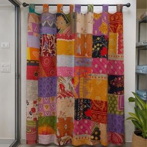 Pode incluir: Uma cortina de patchwork apresentando uma variedade de quadrados de tecido coloridos com diversos padrões florais, geométricos e paisley em tons de vermelho, amarelo, laranja, roxo e verde, suspensa por uma barra de metal escura.