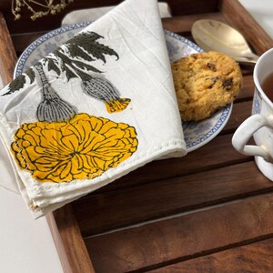 May include: A white cloth napkin featuring a printed design of a large yellow marigold flower with dark green leaves and stems. The napkin has a visible zigzag stitch along the edge and is folded to display the floral pattern clearly.