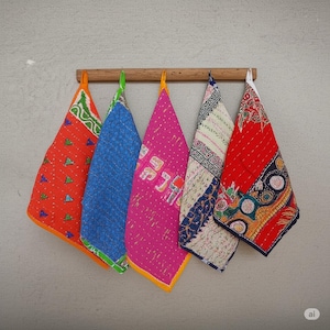 May include: Five colorful, quilted fabric pieces hanging from a wooden rod. The pieces are triangular, with vibrant patterns in red, blue, pink, and white. Each piece has a different design and color scheme, creating a visually appealing display.