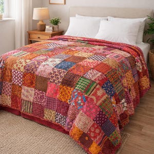Pode incluir: Uma colcha de retalhos com uma mistura vibrante de cores e padrões, incluindo vermelhos, laranjas, rosas e azuis. A colcha está espalhada sobre uma cama com travesseiros brancos, criando uma cena de quarto aconchegante. O design da colcha é feito de muitos quadrados.