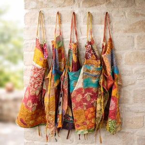 Puede incluir: Una colección de coloridos delantales de patchwork colgados en una pared de ladrillo. Los delantales presentan una variedad de patrones y colores, incluyendo rojo, amarillo y azul. Cada delantal tiene una correa larga para colgar.