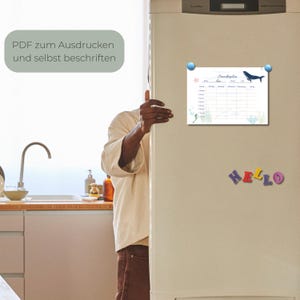 May include: A white magnetic weekly planner with a blue whale illustration is attached to a refrigerator. The planner has a grid layout with days of the week and space for notes. The word "Hello" is spelled out with colorful magnetic letters.