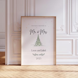 Könnte beinhalten: Ein gerahmter Druck mit dem Text "OUR FIRST CHRISTMAS AS Mr & Mrs" über einer Aquarellillustration eines Weihnachtsbaums. Die Namen "Leon and Juliet" und das Jahr "2025" stehen darunter. Der Rahmen ist goldfarben.