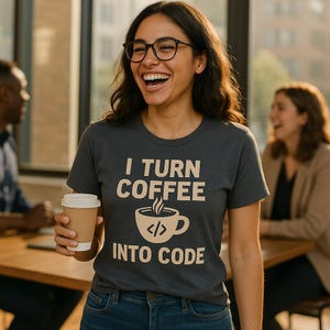 Op de afbeelding: Een vrouw draagt een donkergrijs t-shirt met de tekst "I TURN COFFEE INTO CODE" in lichtbeige. Het ontwerp bevat een koffiekopje met codesymbolen. Ze houdt een bruine papieren koffiebeker vast.