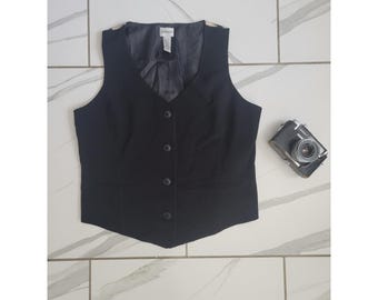 May include: A black vest with a V-neck, button-down front, and two front pockets. The vest has a tailored fit and a dark lining. A vintage camera is next to the vest.