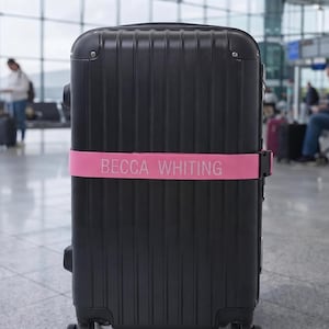 Könnte beinhalten: Ein schwarzer Hartschalenkoffer mit vertikalen Rillen, der horizontal von einem leuchtend pinkfarbenen Kofferband umschlossen ist. Auf dem Band steht in weißer, serifenloser Großschrift der Text BECCA WHITING gedruckt.