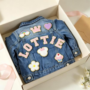 May include: A blue denim jacket featuring various embroidered patches on the back, including a rainbow, a pink crown, a purple heart, a white flower, a cupcake, and a white mouse head shape. The name LOTTIE is spelled out in large pink letters.