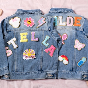 Puede incluir: Dos chaquetas vaqueras azul claro con parches coloridos. Una chaqueta dice "STELLA" y la otra "CHLOE" en letras con lentejuelas. Los parches incluyen una flor, un rayo, una mariposa y un arcoíris.