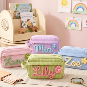 May include: Four pastel-coloured corduroy cosmetic bags. Each bag features an embroidered name and a flower accent. The names are "Mia", "Lily", "Zoe", and "Hannah". The bags are in pink, purple, green, and blue.