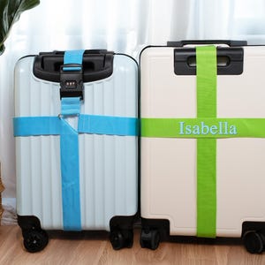 May include: Two wheeled suitcases, one light blue and one cream, are secured with bright straps. The light blue case has a blue strap with a combination lock. The cream case has a green strap with the name "Isabella" printed on it.