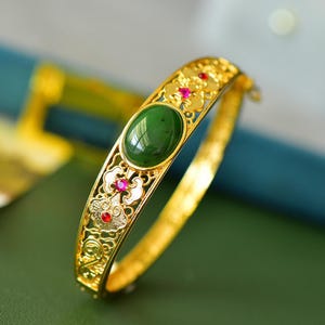 Könnte beinhalten: Ein goldfarbener Klapparmreif mit einem ovalen, dunkelgrünen Stein in der Mitte. Das Metallband zeigt ein kompliziertes, durchbrochenes Filigranmuster, das mit kleinen, runden, leuchtend roten Edelsteinen akzentuiert ist, die im Design eingebettet sind.