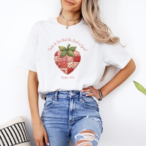 May include: White t-shirt featuring a strawberry design with a patchwork pattern in red, pink, and cream colors. The text "Taste & See that the Lord is Good" and "Psalm 34:8" are printed above and below the strawberry graphic.