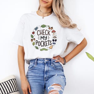 Puede incluir: Camiseta blanca con el texto "CHECK MY POCKETS" en un diseño circular con hojas, insectos y setas. La camiseta se combina con unos vaqueros azules.