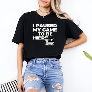 May include: Black t-shirt with the text "I PAUSED MY GAME TO BE HERE" in white, along with a graphic of an airport control tower and airplane. The shirt is paired with blue jeans.