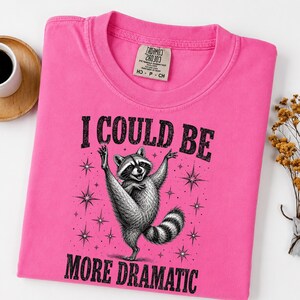 Puede incluir: Una camiseta de color rosa brillante con un gráfico negro de un mapache de pie sobre una pata con los brazos levantados. El texto "I COULD BE MORE DRAMATIC" está impreso en negro sobre y debajo del mapache, rodeado de pequeñas estrellas.