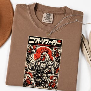May include: A brown crew neck t-shirt featuring a graphic print of a muscular rooster in a martial arts pose. The design includes Japanese text, the words ROUSTEN FIGHTER, and a red sun background, framed by four smaller rooster head panels.
