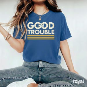May include: Royal blue t-shirt with the words "GOOD TROUBLE" in a cream-colored font, with two horizontal gold stripes underneath. The shirt is a crew neck and has short sleeves.