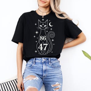 Peut inclure: T-shirt noir avec un motif blanc de chat, d'étoiles, d'un croissant de lune et des chiffres 86 et 47. Le design du chat est fantaisiste. Le t-shirt est porté avec un jean bleu.