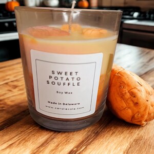 May include: A glass candle with a label reading "SWEET POTATO SOUFFLE" and "Soy Wax." The candle has orange and yellow hues. A whole sweet potato is placed beside the candle on a wooden surface.