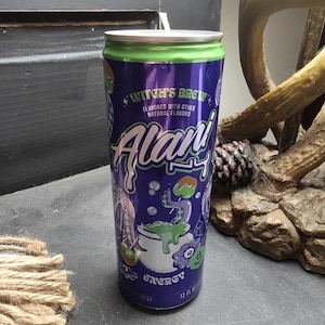 Peut inclure: Une canette de boisson énergisante Alani Nu violette et verte étiquetée Witch's Brew, avec un graphique représentant un chaudron bouillonnant, un tentacule et des pommes d'amour. La canette a un dessus argenté, contient 355 ml et porte le texte Flavored with other natural flavors.