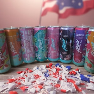 May include: A selection of Alani Nu Energy drink cans with diverse, vibrant designs and flavours, such as cherry and tropical fruit. The cans are displayed on a wooden surface with red, white, and blue decorations, with a flag in the background.