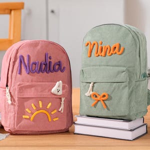 Könnte beinhalten: Zwei Cord-Rucksäcke, einer rosa mit "Nadia" in lila Schrift und einem Sonnendesign, der andere grün mit "Nina" in orangefarbener Schrift und einer Schleife. Beide haben Vordertaschen und stehen auf einer Holzoberfläche.