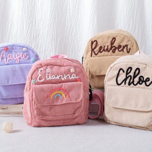May include: Four corduroy backpacks in pastel colors: lavender, pink, tan, and cream. Each backpack has a front pocket and is personalized with a name embroidered in a contrasting color. The pink backpack features a rainbow design.