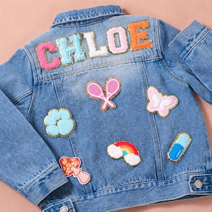 May include: A light blue denim jacket featuring the name CHLOE in large, colorful, glitter-bordered letters across the back. The jacket is decorated with various embroidered patches, including tennis rackets, a butterfly, a rainbow, and a mushroom.