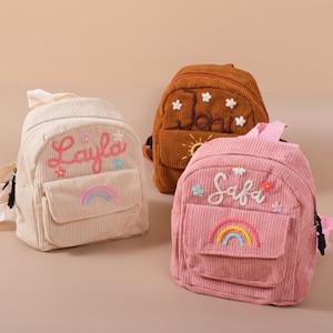 May include: Three corduroy backpacks in cream, brown, and pink. Each backpack has a front pocket and is embroidered with a name and floral and rainbow designs. The cream backpack says "Layla", the brown backpack says "Joni", and the pink backpack says "Safa".