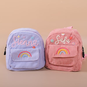 May include: Two small corduroy backpacks, one lavender and one pink. Each features a rainbow, embroidered name, and floral accents. The lavender backpack says "Sienna" and the pink backpack says "Safa". Both have a front pocket and black zippers.