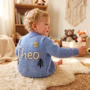 May include: A light blue long-sleeve knit jumper featuring the name Theo embroidered in yellow yarn on the back. The design includes a yellow crescent moon above the name and two dark green pine trees embroidered below the name.