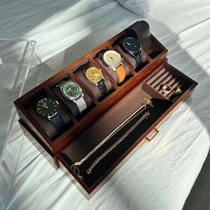 May include: A wooden watch and jewelry organizer with multiple compartments. The top section holds several watches with different colored bands. The bottom drawer contains gold necklaces and rings. The text "FahminStudio" is visible in the image.