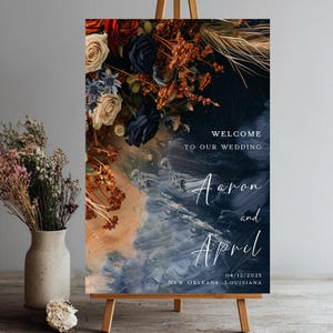 May include: A wooden easel displays a wedding welcome sign. The sign features floral arrangements in shades of red, navy, and cream. Text on the sign reads "Welcome to our wedding" and the names "Aaron and April" with the date and location.