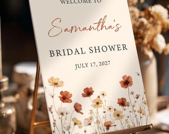 May include: A cream-colored sign on a wooden easel, with the words "WELCOME TO" at the top, followed by "Samantha's" in a cursive font, and "BRIDAL SHOWER" and "JULY 17, 2027" below. The bottom features a floral border with orange, yellow, and white flowers.