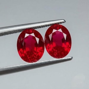 May include: Two oval-cut, vibrant red gemstones held by silver-toned metal tweezers. The stones feature a faceted surface that reflects light, showcasing a deep crimson color and a clear, polished appearance against a neutral background.