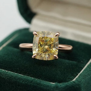 May include: A ring featuring a large, cushion-cut yellow gemstone set in a polished rose gold band. The stone displays a multifaceted surface with warm, golden hues, held securely by four prongs atop a smooth, rounded metal shank.