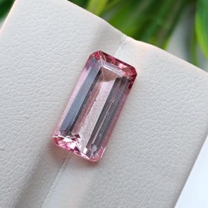 Può includere: Una gemma rettangolare e sfaccettata con una tonalità rosa. La gemma ha un aspetto chiaro e trasparente ed è posta su una superficie bianca. Il taglio e la purezza della gemma sono evidenti, mostrando la sua struttura interna e il colore.