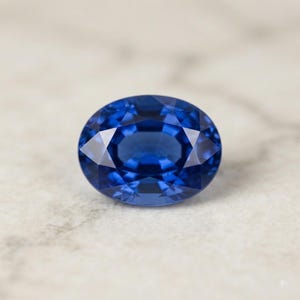 May include: A vibrant, deep blue gemstone cut into a classic oval shape. The stone features a faceted surface with a symmetrical arrangement of triangular and kite-shaped facets that reflect light, creating a brilliant, sparkling appearance.