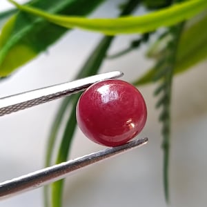 May include: A polished, round, deep red gemstone held by silver tweezers. The gem has a smooth, reflective surface, and is set against a blurred background of green foliage and a white surface. The gemstone appears to be a ruby.