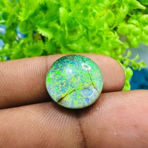 May include: A round, iridescent opal gemstone with a cracked pattern. The opal displays a mix of green, blue, and yellow hues, with a dark base. The gemstone is held in a hand, with a blurred green background.