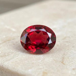 May include: A deep red, oval-cut gemstone with a faceted surface that reflects light. The stone displays a rich, saturated crimson hue and a symmetrical arrangement of triangular and kite-shaped facets across its top, creating a brilliant appearance.