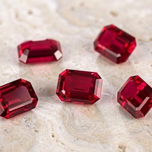 May include: Five loose, vibrant red gemstones with an emerald cut, featuring rectangular shapes and faceted surfaces that reflect light. These deep red stones are arranged in a scattered pattern, showcasing their clear, polished, and transparent appearance.