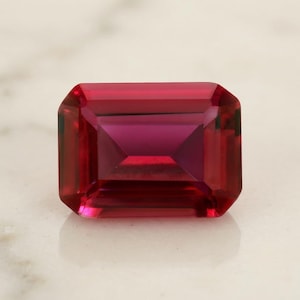 May include: A vibrant, deep red gemstone cut into a rectangular emerald shape. The stone features precise, clean facets that create a symmetrical geometric pattern, reflecting light with a rich, saturated crimson hue across its polished surface.