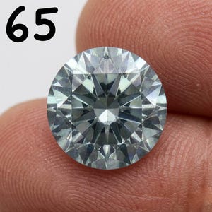 Könnte beinhalten: Ein runder Edelstein im Brillantschliff mit einem kühlen, eisblauen bis grauen Farbton und einer facettierten Oberfläche, die das Licht reflektiert. Die Zahl 65 ist in Schwarz in der oberen linken Ecke des Bildes aufgedruckt.