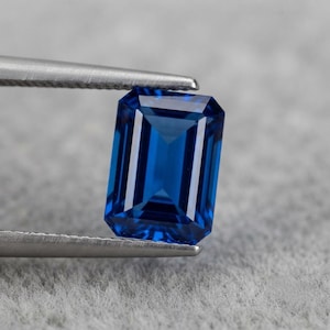 May include: A vibrant blue gemstone with an emerald cut, featuring a rectangular shape and faceted surfaces that reflect light. The gemstone is held securely by a pair of silver-toned metal tweezers, showcasing its deep colour and clarity.