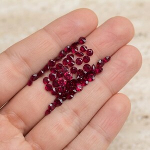 Könnte beinhalten: Eine Sammlung kleiner, runder, facettierter loser Edelsteine in einer tiefen, leuchtend roten Farbe. Diese Steine weisen einen Brillantschliff mit mehreren reflektierenden Oberflächen auf und zeigen einen einheitlichen karmesinroten Farbton.