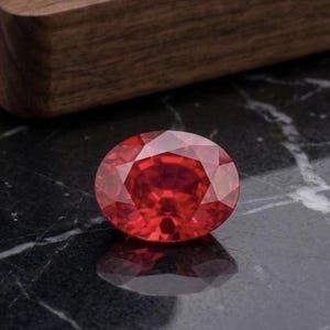 Puede incluir: Una gema roja de corte ovalado con una superficie facetada que refleja la luz. La piedra muestra un tono carmesí profundo y saturado con múltiples facetas triangulares y en forma de cometa dispuestas en un patrón simétrico superior.