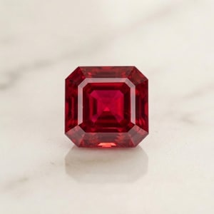 Op de afbeelding: Een levendig rode edelsteen met een vierkante trapslijpvorm, voorzien van meerdere concentrische facetten die een geometrisch patroon vormen. De steen vertoont diepe karmozijnrode tinten en scherpe, heldere randen die het licht reflecteren.