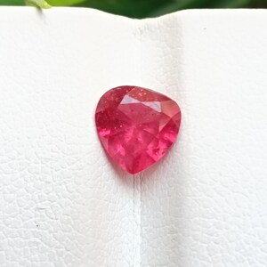 May include: A faceted, heart-shaped, ruby-red gemstone. The gem is set against a white background, with a hint of green foliage in the upper left corner. The stone's facets catch the light, creating a sparkling effect.