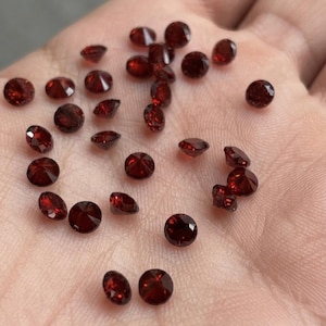 Puede incluir: Una colección de pequeñas piedras preciosas de color rojo oscuro, redondas y facetadas. Cada piedra presenta un corte brillante con múltiples facetas triangulares que reflejan la luz, creando un profundo brillo carmesí en la superficie.